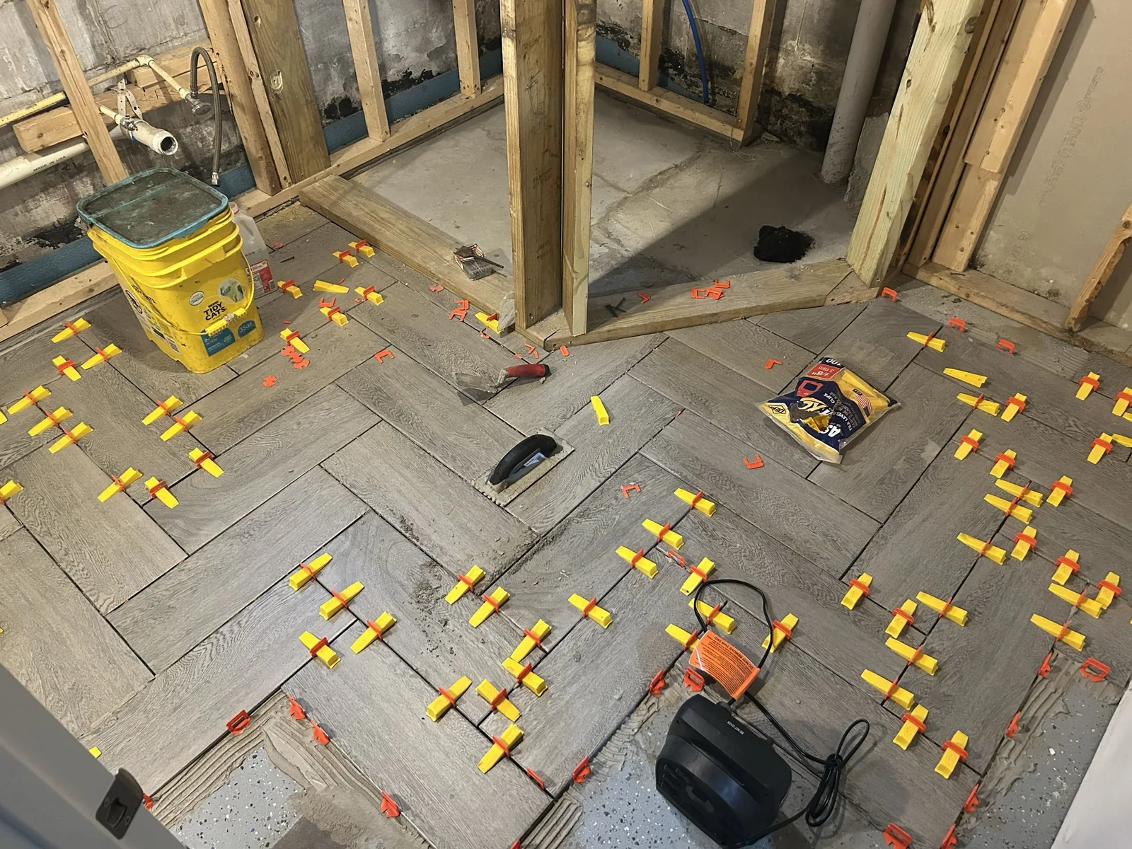 Wood-look herringbone tile floor going down with spacers beside new bathroom framing