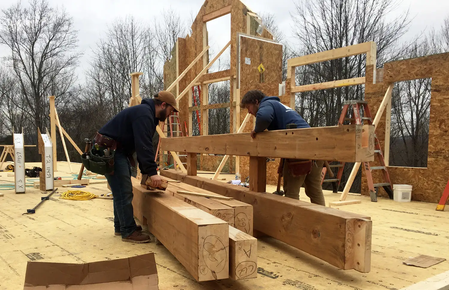 Two carpenters working heavy timber beams on the deck of a house under construction